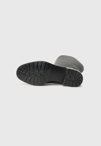 Bota de borracha preta com uma sola texturizada que apresenta ranhuras profundas para aderência. O interior da bota é liso, e a forma geral é cilíndrica.