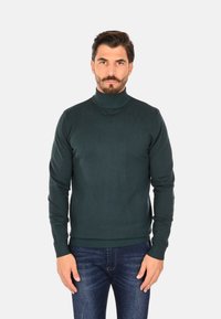 Maglione a collo alto verde scuro in maglia a coste. Linea aderente con maniche lunghe e collo alto. Abbinato a jeans blu.