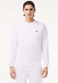 Jeune homme aux cheveux foncés portant un sweat-shirt blanc uni et un pantalon blanc assorti, avec un petit logo crocodile vert sur la poitrine.