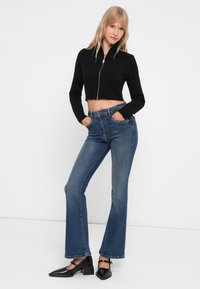 Svart kort zip-up genser parret med blå flagrende jeans og svarte blokkhælede sko. Glatt stoff og tettsittende design.