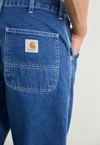 Denim jeans with a blue hue, featuring a back pocket with a white label bearing a yellow logo. Stitched detailing along the edges.