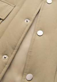 Primer plano de una chaqueta de tela beige con costuras visibles y botones automáticos plateados a lo largo de la apertura frontal.