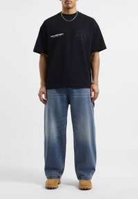 Camiseta negra de gran tamaño con gráfico en el pecho, combinada con jeans de pierna ancha en azul claro y zapatos beige, con un corte relajado y un ligero desgaste.
