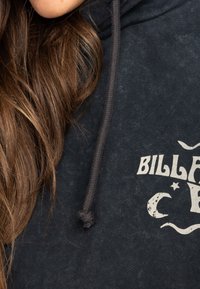 Detailní záběr na dlouhé hnědé vlasy přes tmavou mikinu s kapucí a částečně bílým textem "Billabong."