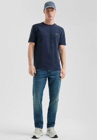 Jonge man in een marineblauwe t-shirt, blauwe spijkerbroek, grijze pet en witte sneakers, staand tegen een effen lichtgrijze achtergrond.