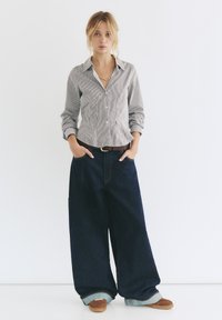 Camicia a righe con bottoni in grigio chiaro e bianco, abbinata a pantaloni in denim blu scuro a gamba larga. Scarpe marroni con dettaglio del risvolto.