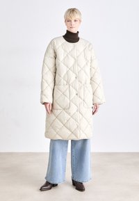 Casaco longo bege acolchoado com grandes costuras em diamante, gola redonda e botões frontais. Combinado com jeans azuis de perna larga e botas castanhas.