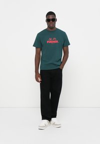 Dark green cotton t-shirt with red "FORMER." text and graphic, paired with black pants and white sneakers. Features short sleeves and a crew neck.