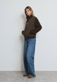 Veste zippée marron avec un devant en daim et des manches côtelées, associée à un jean large bleu clair et des chaussures marron, sur un fond uni.