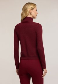 Burgunderfarbener Rollkragenpullover mit enganliegendem Design, geripptem Saum und Bündchen, aus weichem, strukturiertem Material. Getragen mit passenden Hosen.