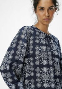 Blusa azul marino con patrones florales blancos, cuello redondo y detalle plisado en la parte frontal. Mangas largas con ajuste relajado. Tejido texturizado.
