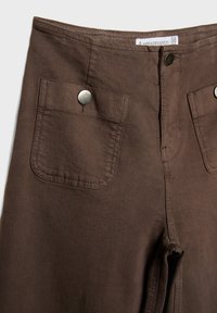 Pantalon marron taille haute avec poches avant fermées par des boutons-pression métalliques, coutures apparentes et étiquette taille 38 à l'intérieur de la ceinture.