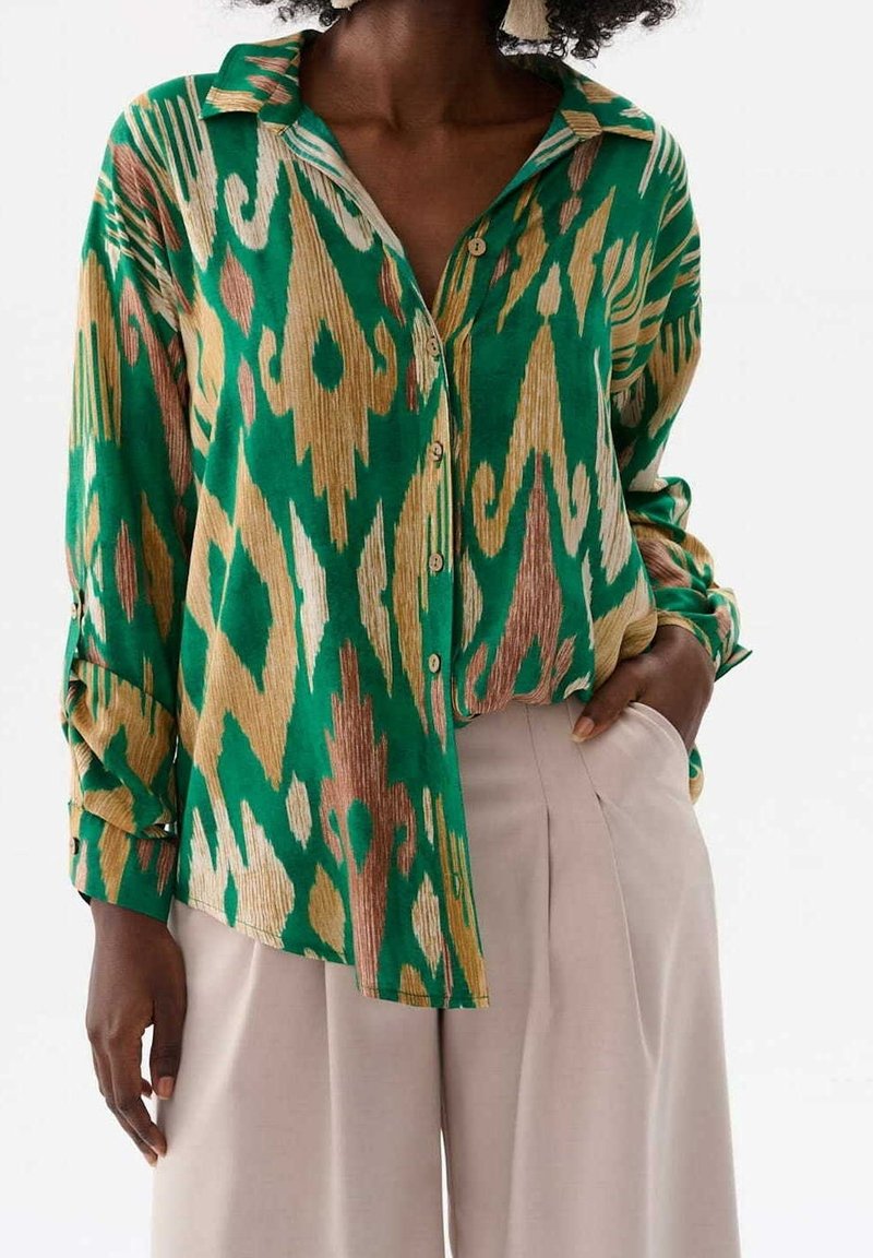 Personne à la peau foncée portant une chemise boutonnée à motifs verts, beige et marron avec un pantalon beige taille haute, main dans la poche.