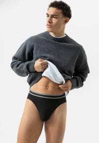 Grauer Strickpullover mit gerippten Bündchen, kombiniert mit schwarzen gerippten Slips mit einem kontrastierenden grauen Bund. Das Model posiert vor einem weißen Hintergrund.