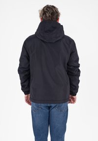 Uomo con una giacca nera con cappuccio e jeans blu, in piedi di spalle su uno sfondo bianco.