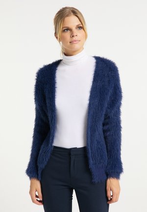 Donna bionda che indossa un dolcevita bianco e un cardigan aperto davanti blu navy morbido, in piedi davanti a uno sfondo chiaro e semplice.