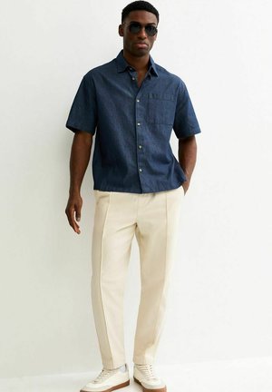 Mężczyzna w czarnych okularach przeciwsłonecznych, ciemnoniebieskiej koszuli na krótki rękaw z guzikami, jasnobeżowych spodniach i białych tenisówkach, stojący z jedną ręką w kieszeni.