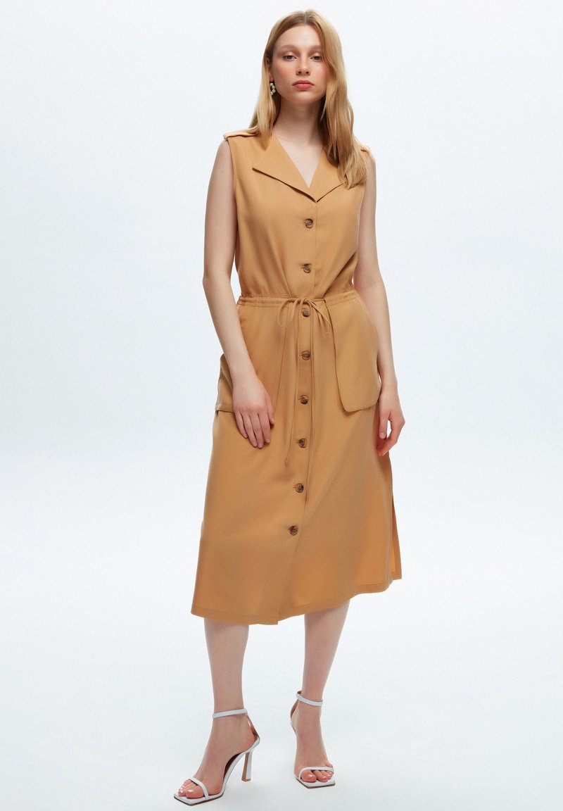 Abito midi beige con colletto, bottoni frontali e vita sottile. Design senza maniche con tasche laterali. Tessuto morbido; leggero lucido.