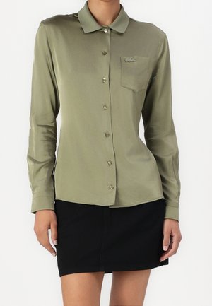 Kobieta ubrana w oliwkową koszulę na guziki z kieszenią na piersi i czarną spódnicę, stojąca na tle jednolitego białego tła.