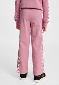 Roze sweatpants gemaakt van zachte stof, met witte chevronstrepen langs de zijkant en een relaxte pasvorm. Zwarte schoeisel zichtbaar aan de onderkant.