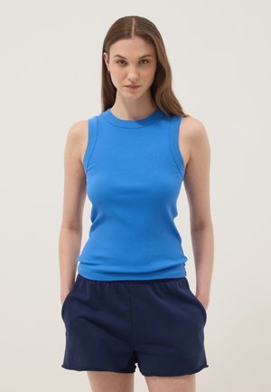 Junge Frau mit langen Haaren, die ein blaues ärmelloses Ripp-Tanktop und dunkelblaue Shorts trägt, steht mit den Händen in den Taschen vor einem einfarbigen Hintergrund.