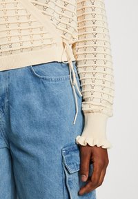 Knit sweater in cream with a textured pattern, drawstring waist, and ruffled cuffs; paired with blue denim cargo pants featuring a side pocket.