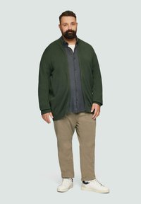 Grüner Cardigan, darunter ein graues, knopfbares Shirt, beige Hose und weiße Sneaker. Weicher Stoff mit lässiger Passform. Minimalistisches Design.
