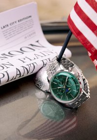 Ceas Timex cu cadran verde, brățară argintie din oțel inoxidabil, carcasă rotundă și trei subcadrane. În apropiere se află un ziar și un steag.