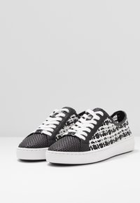 Baskets tissées noires et blanches basses avec lacets blancs et semelles épaisses blanches, placées côte à côte en angle sur une surface blanche.