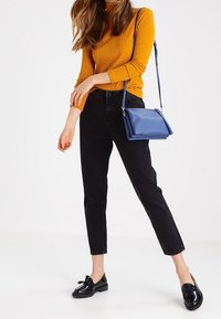 Žebrovaný hořčicový svetr, černé slim-fit džíny a černé mokasíny. Na tenkém popruhu je nošena modrá křížová taška.