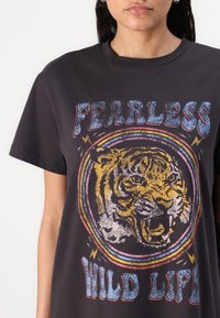 Melnas kokvilnas T-krekls ar tīģera grafiku un uzrakstu "NEBAIDĪGA SAVVAĻA DZĪVE", krāsainiem akcentiem un apaļu kakla izgriezumu.