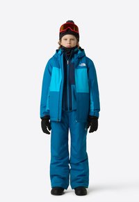 Modrý lyžařský úbor sestávající z bundy s kapucí a kalhot, vyrobený z odolného materiálu proti povětrnostním vlivům. Obsahuje černé rukavice a brýle s červeným tónem.