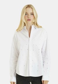 Camicia bianca con bottoni, dal taglio rilassato, caratterizzata da decorazioni metalliche sparse e colletto classico. Maniche lunghe con polsini abbottonati.