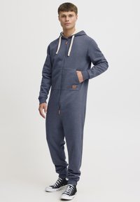 Navy blue zip-up hoodie jumpsuit with a cream-lined hood, front pocket, and a leather label accent; features elastic cuffs and relaxed fit.