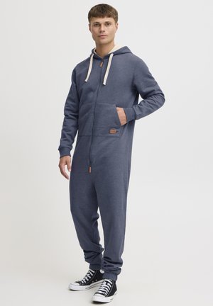 Navy blue zip-up hoodie jumpsuit with a cream-lined hood, front pocket, and a leather label accent; features elastic cuffs and relaxed fit.