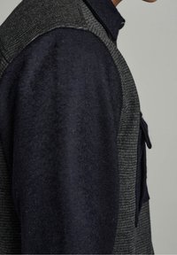 Gilet misto lana con sezioni in navy scuro e grigio a spina di pesce, caratterizzato da una finitura testurizzata e una tasca sul petto sul lato sinistro.