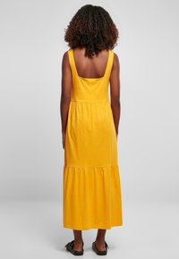 Żółta, bezrękawna sukienka maxi z kwadratowym dekoltem na plecach, z falbanami na dole i gładką bawełnianą fakturą. Noszona z płaskimi sandałami.