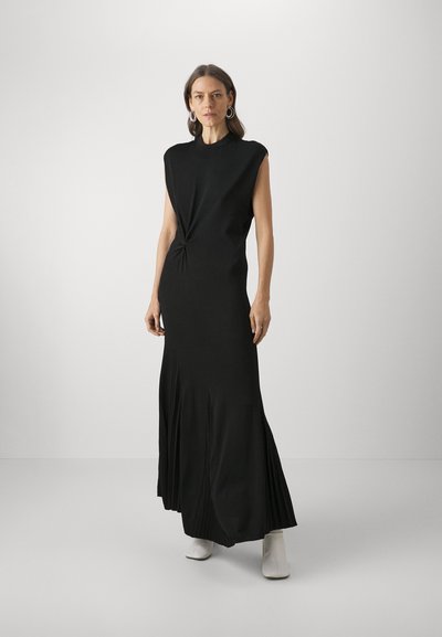 Robe maxi sans manches noire avec un corsage ajusté, détail noué à la taille et ourlet plissé, fabriquée en tissu doux. Bottines blanches.