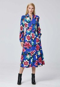 Femme debout dans une robe midi bleue à fleurs avec des manches longues et des bottines noires, posant avec une main dans la poche devant un fond uni.