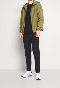 Olivgrön lättviktsjacka med huva, dragkedja och elastiska ärmslut, matchad med svarta randiga byxor och vita sneakers.