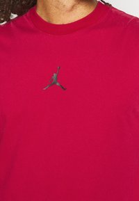 Červené bavlněné tričko s malým černým logem Jumpman na hrudi, kulatým výstřihem a krátkými rukávy. Hladká textura.