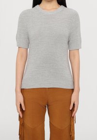 Pull tricoté gris à manches courtes avec une texture côtelée et un col rond, stylé avec un pantalon en daim marron orné de franges.