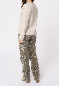 Femme portant un pull beige en tricot et un pantalon taille haute à imprimé léopard, debout de dos sur un fond clair uni.