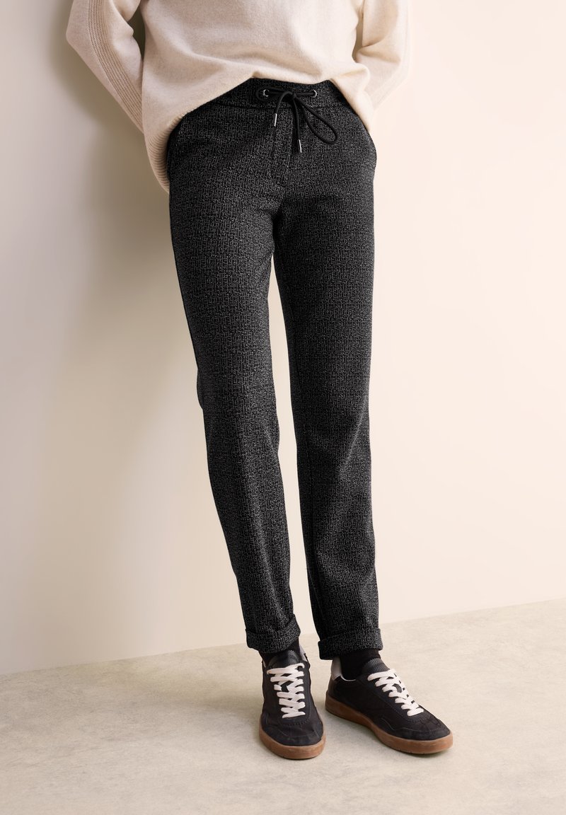 Pantalons de jogging noirs à motif avec une taille élastique et un cordon de serrage, associés à des baskets noires avec des lacets blancs et une semelle marron.