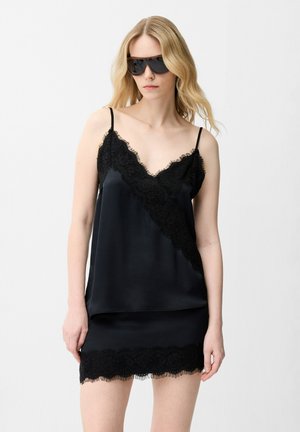 Blonde vrouw die grote zwarte zonnebrillen draagt, een zwart satijnen hemdje met kanten rand en een bijpassende zwarte rok met kanten rand tegen een witte achtergrond.