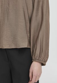 Blusa in tessuto leggero marrone con maniche lunghe e elasticizzate e finitura testurizzata. L'orlo è diritto, completato da un design semplice.
