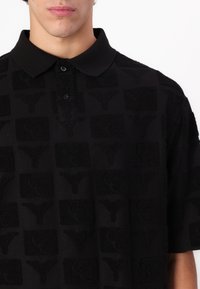 Svart polo-skjorte med et teksturert mønster av firkanter med logodetaljer, en knappestolpe og en klassisk krage. Laget av mykt stoff.