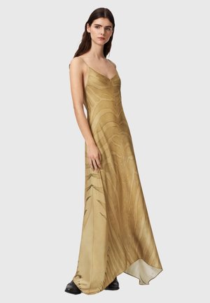 Femme portant une longue robe nuisette dorée à fines bretelles avec des motifs subtils façon zèbre, associée à des chaussures noires, debout devant un fond uni.