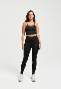 Un crop top de sport noir et un legging taille haute, tous deux présentant une texture lisse. La tenue inclut un branding subtil et est associée à des baskets blanches.