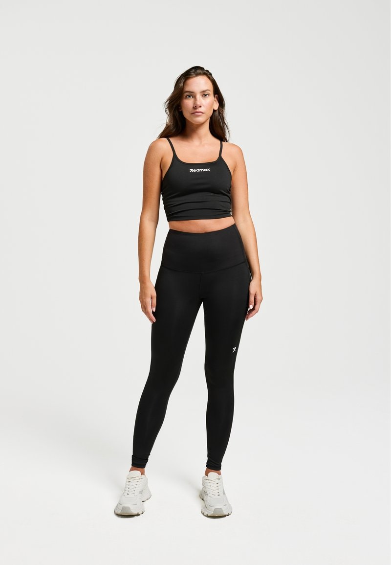 Un crop top de sport noir et un legging taille haute, tous deux présentant une texture lisse. La tenue inclut un branding subtil et est associée à des baskets blanches.